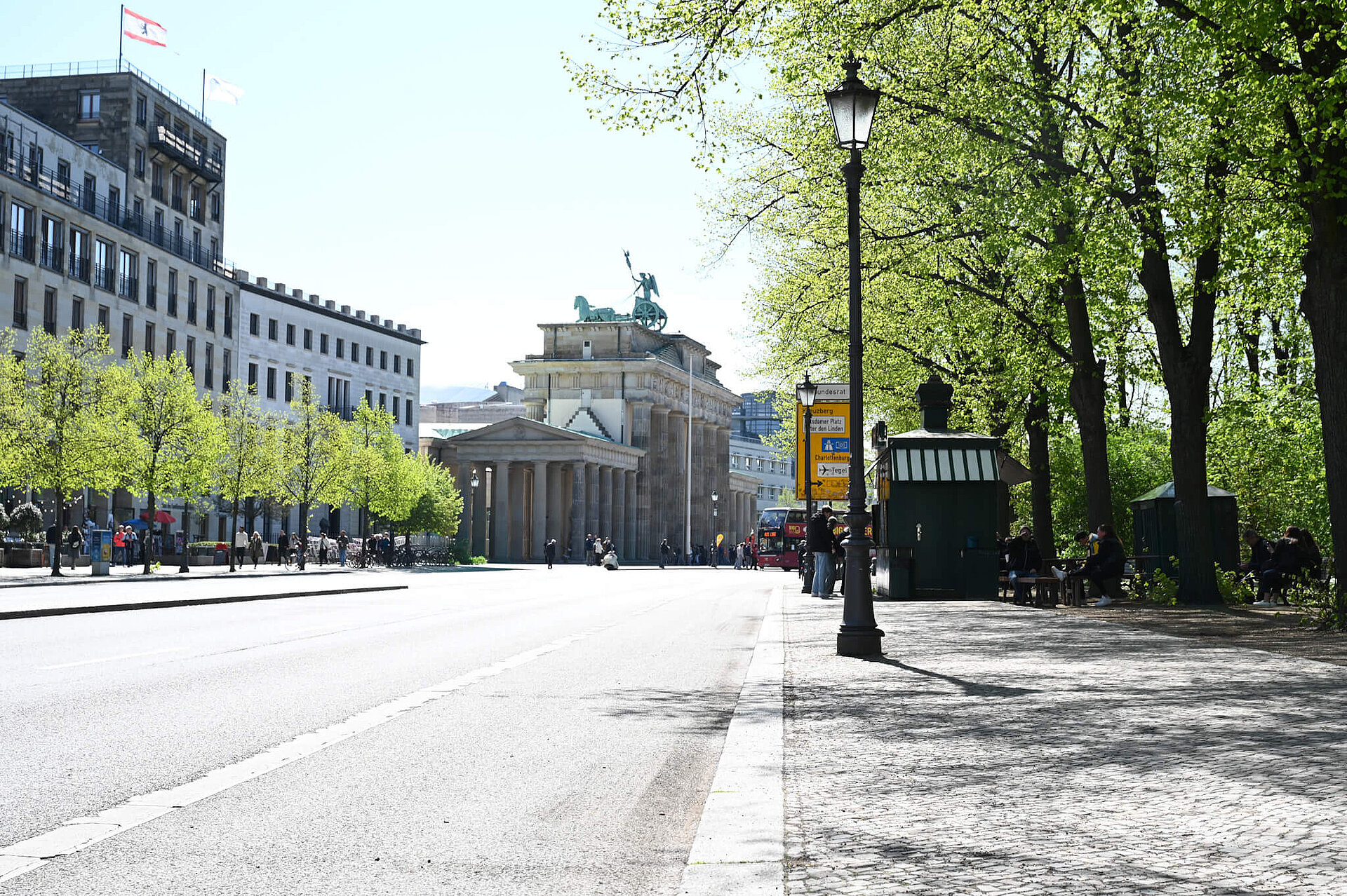 Blick entlang der Straße auf das Brandenburger Tor. Grüne Bäume und ein Zeitungskiosk am Straßenrand.