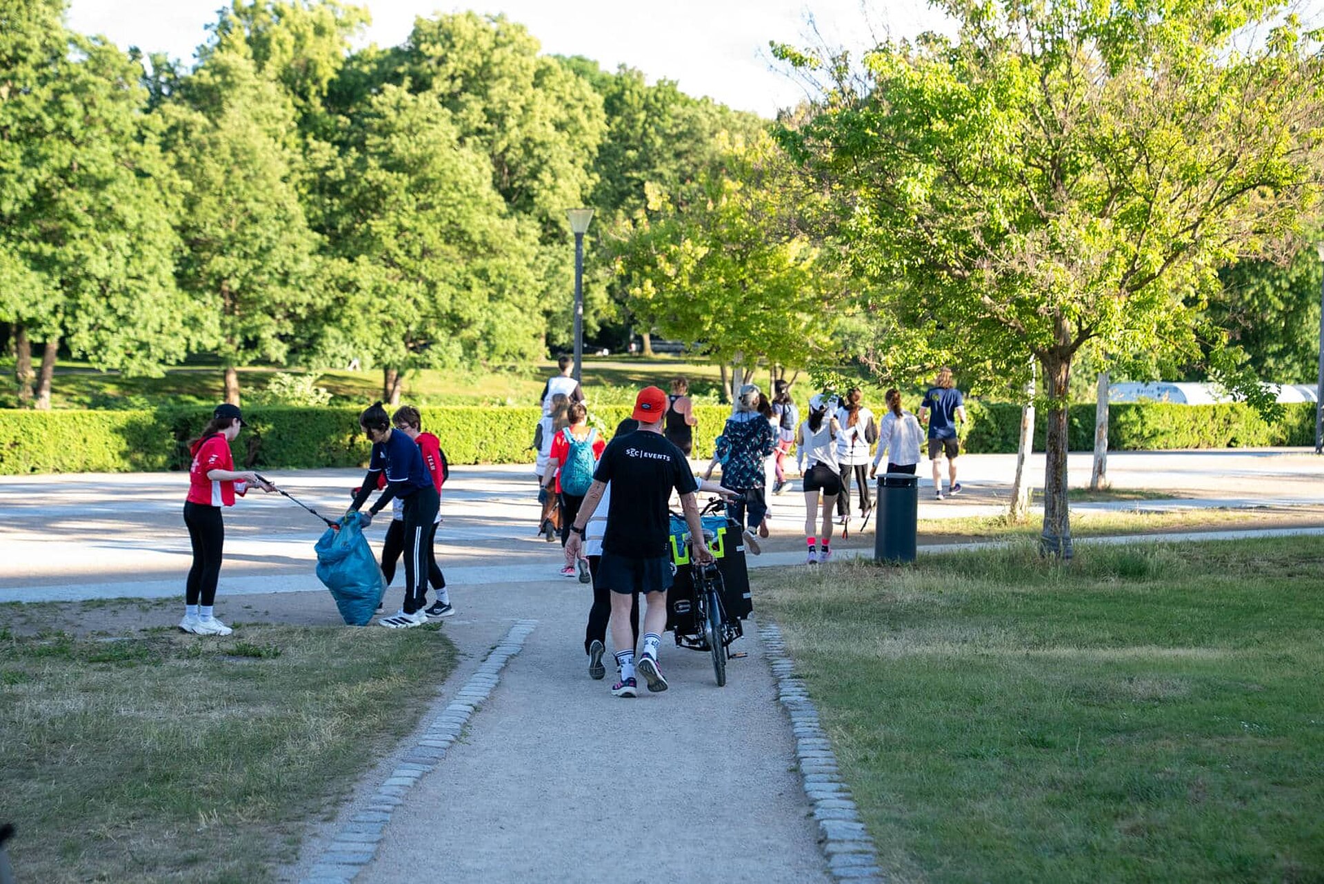 Etwa 15 Teilnehmende sammeln joggend Müll mit Müllgreifern. Hinter der Gruppe wird ein Lastenfahrrad geschoben, das einen blauen Müllsack zu enthalten scheint.