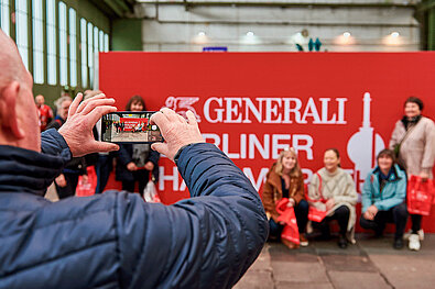 GENERALI BERLIN HALF MARATHON 2023: older man takes a cell phone photo of a group of people in front of the event logo © SCC EVENTS / Steffen Hartz