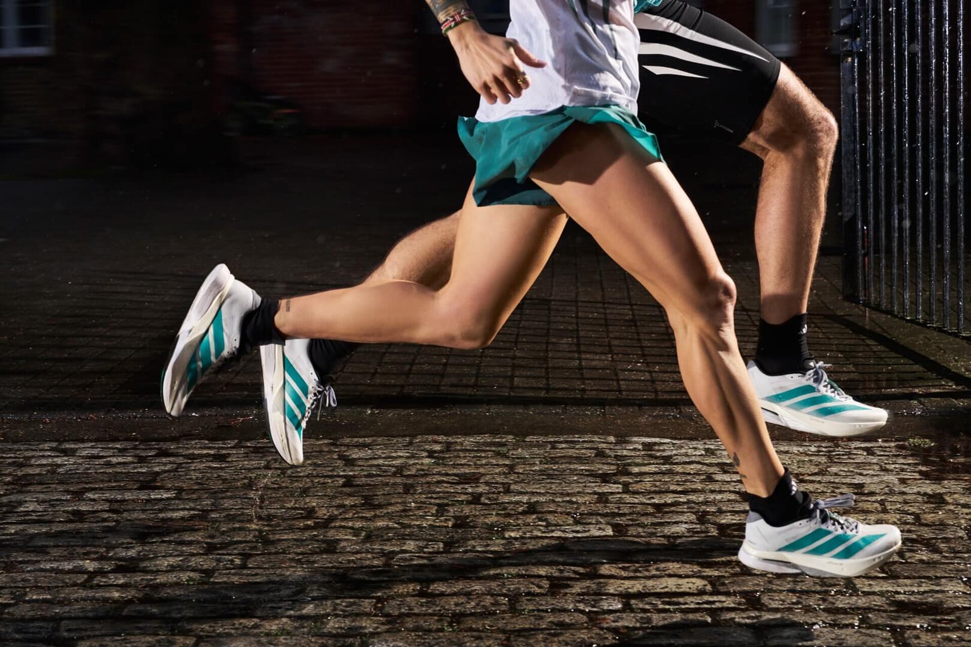 Zwei Läufer:innen in weißen adidas-Schuhen mit grünen Streifen rennen über Pflasterstein. Zu sehen sind nur die Beine.