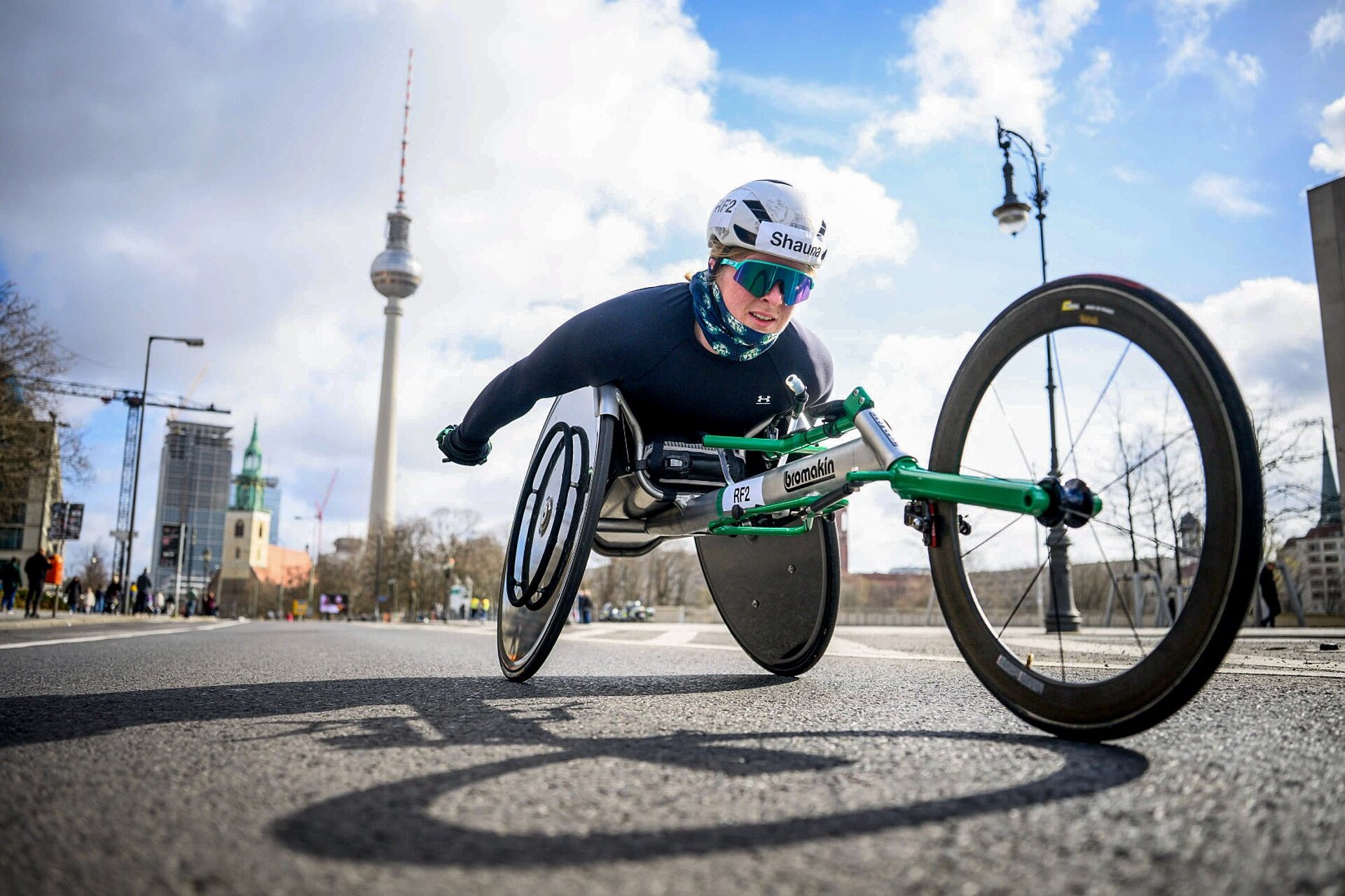 Shauna Bocquet, wearing sunglasses, drives her racing wheelchair right past the camera. Shot from a low-angle perspective.