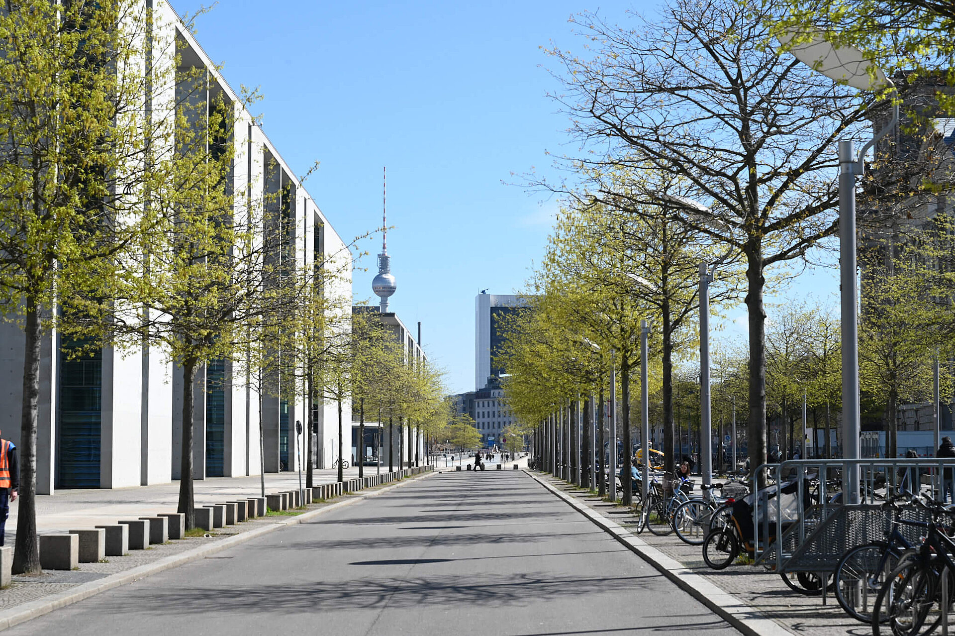 Allee gesäumt von jungen Bäumen. Links das Paul-Löbe-Haus, rechts eine kleine Ecke des Reichstagsgebäudes erkennbar. Im Hintergrund der Fernsehturm.
