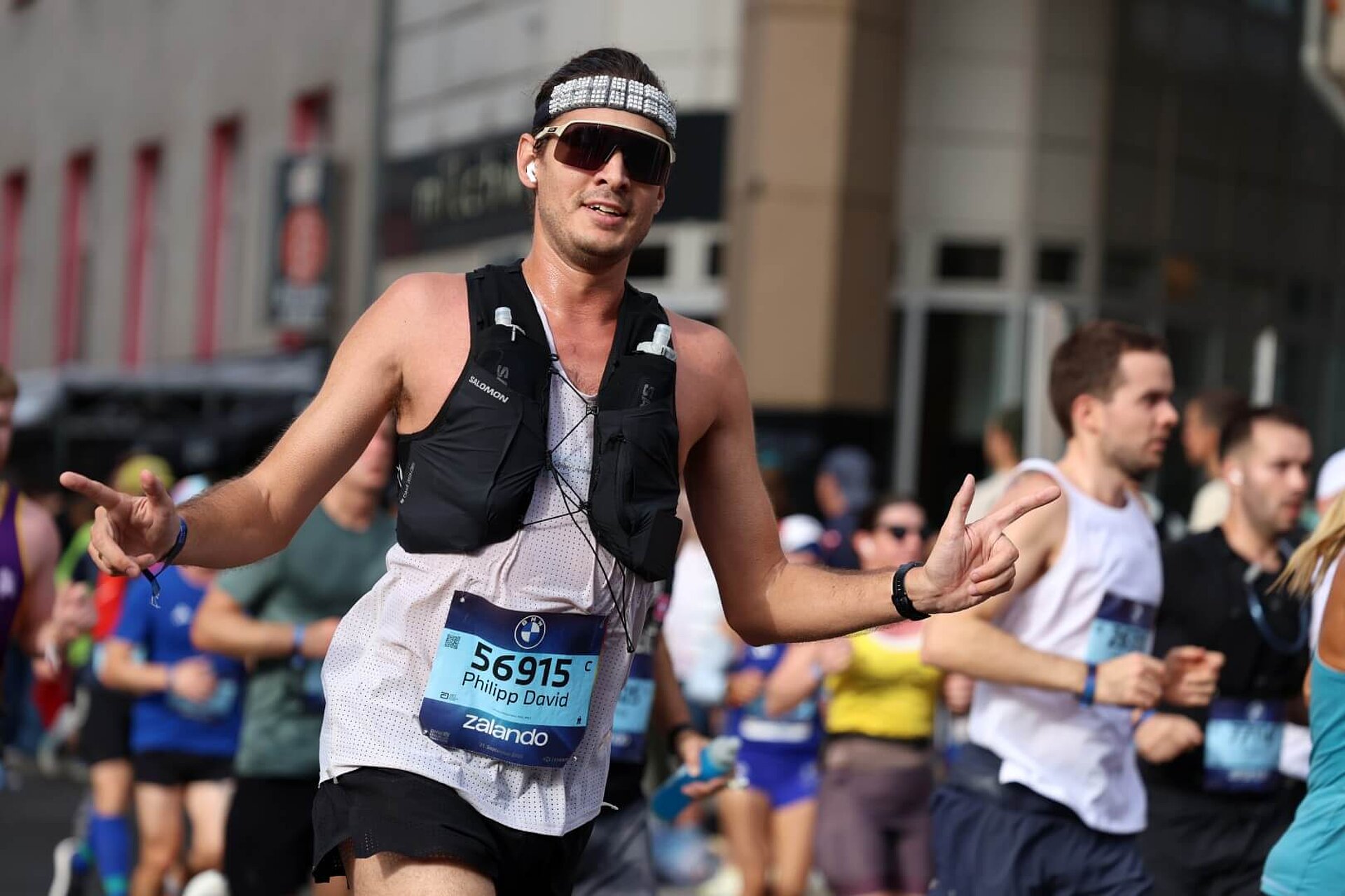 A runner wearing a hydration vest, with the tops of two soft flasks sticking out of it. He has his arms raised halfway in a greeting gesture.