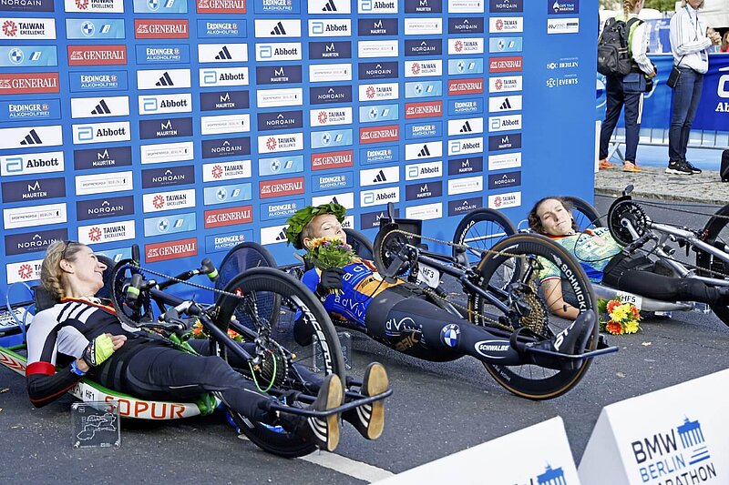 Handbike Siegerinnen von 2023 auf dem Podest in ihren Rädern. Julia Dierkesman mit Lorbeerrkranz auf dem Kopf.