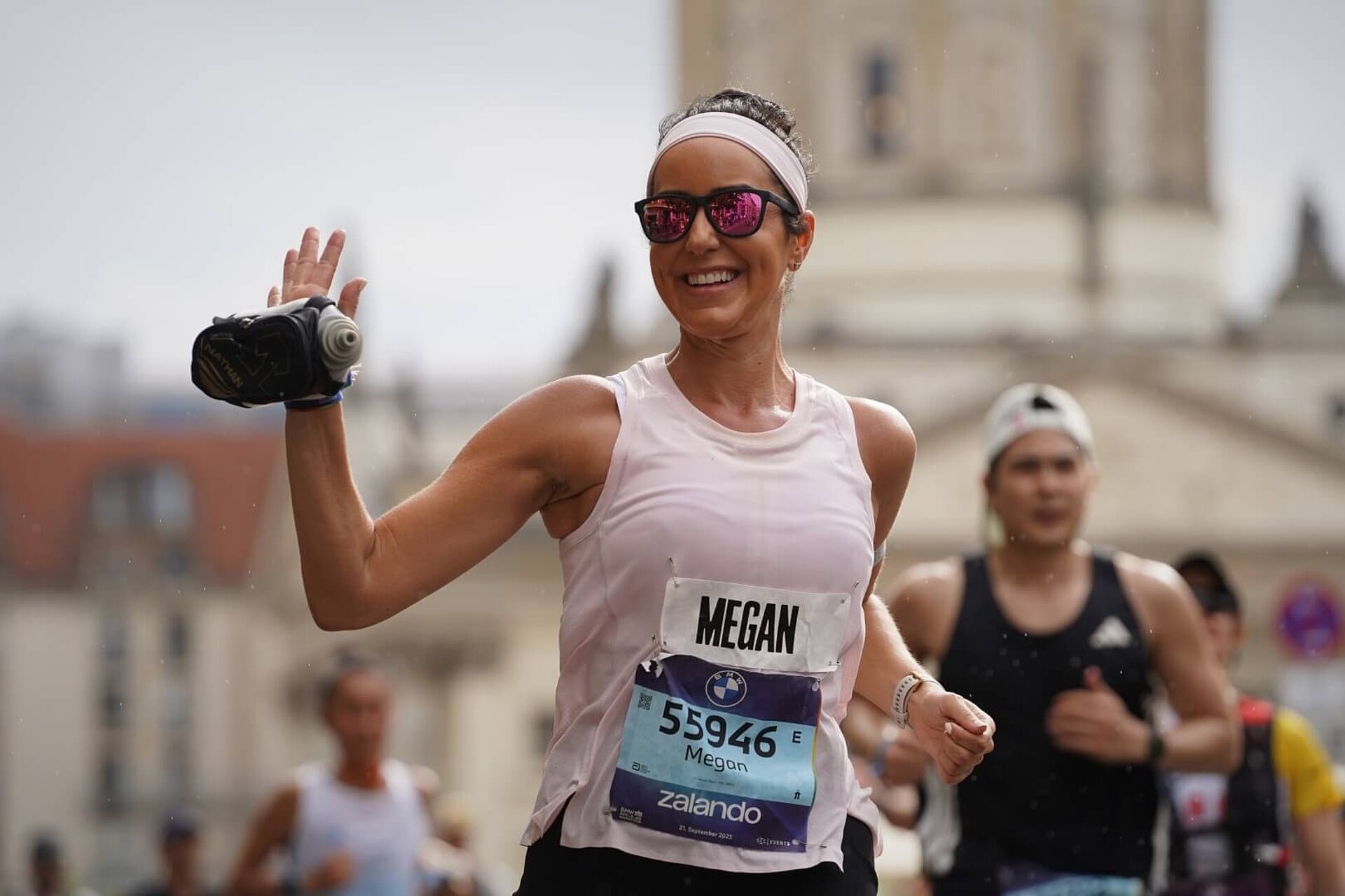 A smiling runner waves with one hand, to which a bottle is attached.