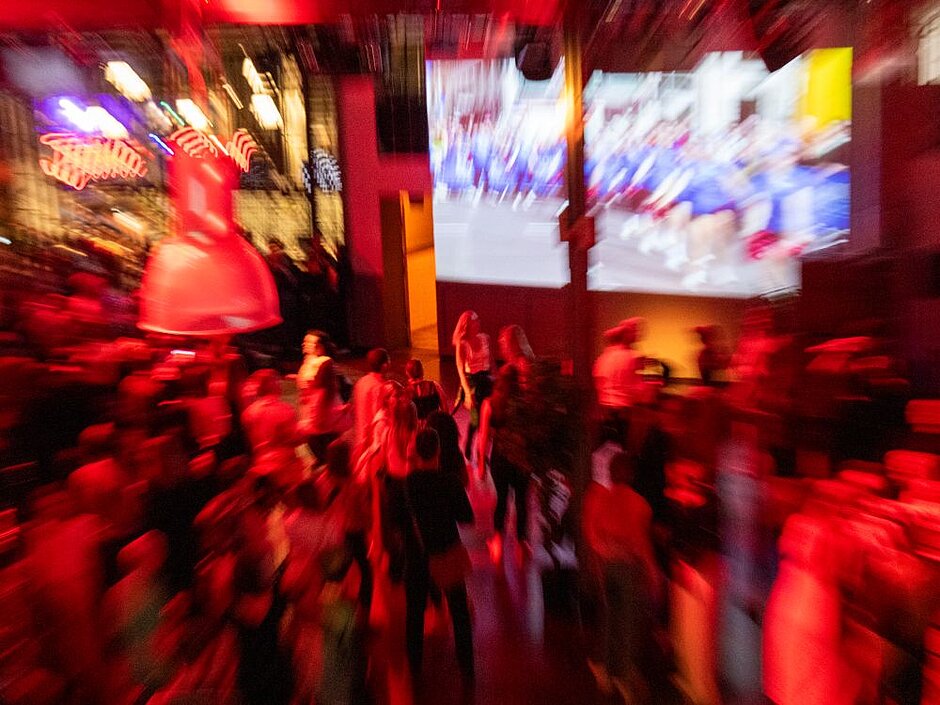 Dance floor bathed in red light in front of a screen that appears to be showing a race. Blurred vignette suggests energetic movement.