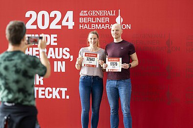 GENERALI BERLIN HALF MARATHON Inline Skating 2024: Couple of participants hold their race numbers up to the camera in front of the GENERALI photo wall © SCC EVENTS / Jean-Marc Wiesner