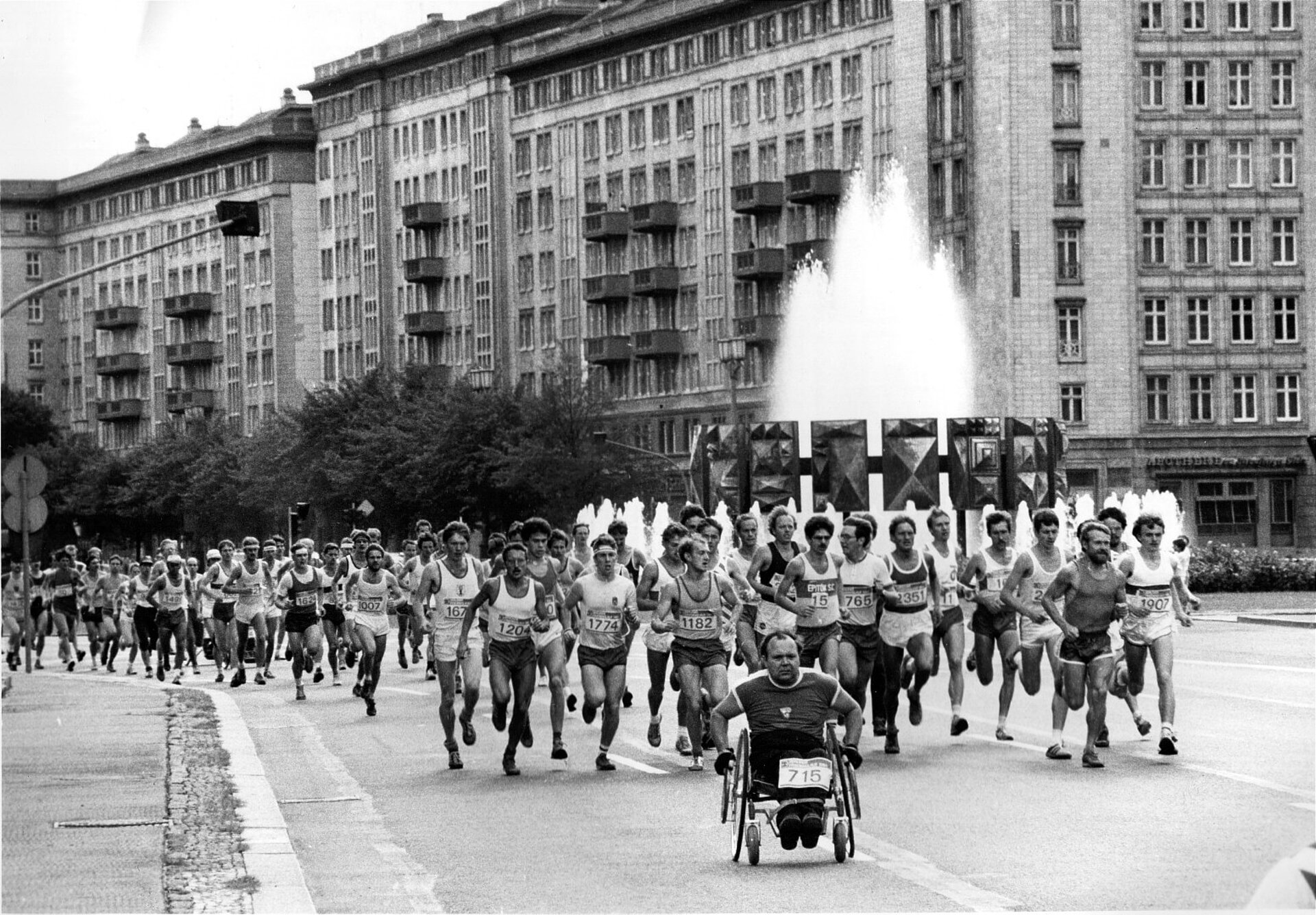 Rollstuhlfahrer vor Läufer:innenfeld. Im Hintergrund ein sprudelnder Springbrunnen.