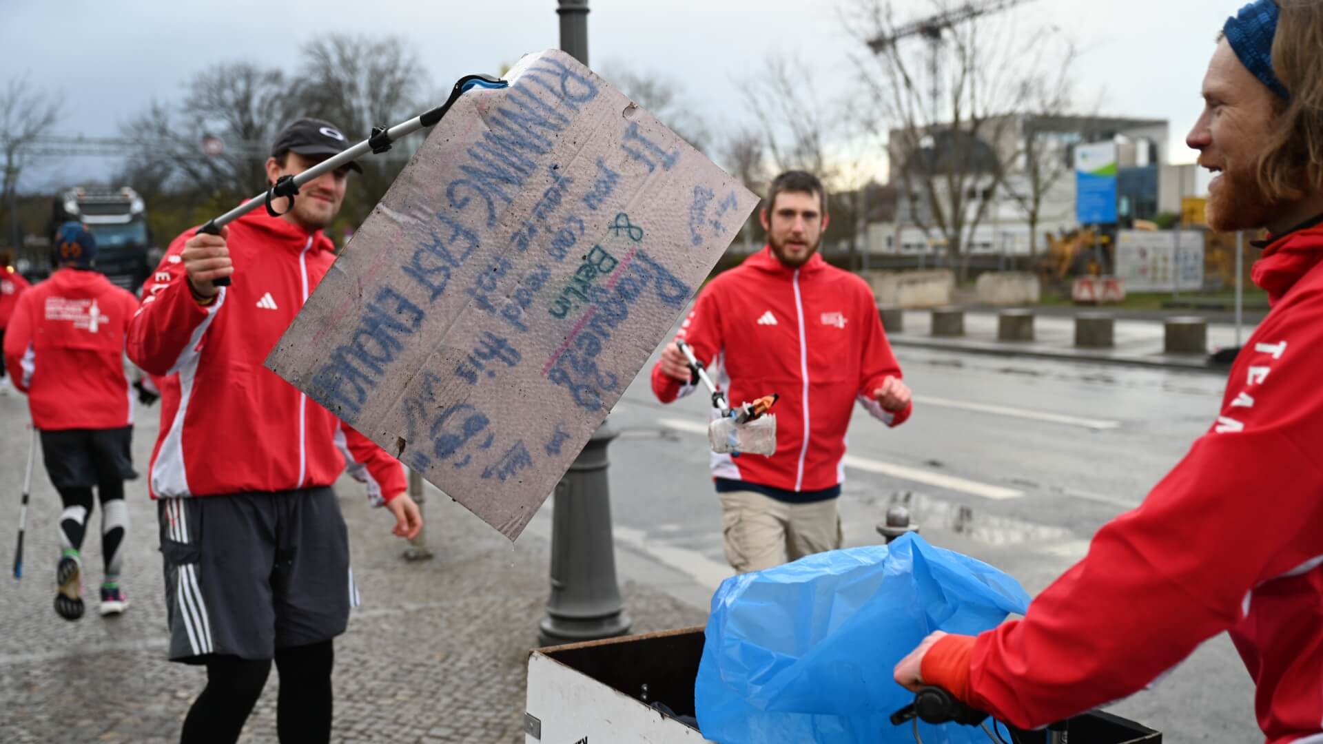 Lächelnder Teilnehmer bringt nasses Cheering Sign des Rennens mit Müllgreifer zum Lastenrad mit Müllsack. Anderer Teilnehmer bringt einen Plastikbecher.